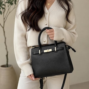 May include: A black leather handbag with gold-colored hardware. The bag has a structured shape with a top handle and a detachable shoulder strap. The bag is held by a person wearing a cream-colored cardigan and white pants.