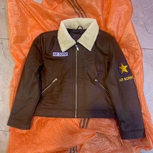 K&ouml;nnte beinhalten: Eine braune Jacke mit einem cremefarbenen Webpelzkragen und einem durchgehenden Frontrei&szlig;verschluss. Auf der Brust befindet sich ein Aufn&auml;her mit dem Text AIR BORNE, und am linken &Auml;rmel ist ein gelbes Sternemblem &uuml;ber dem Text AIR BORNE zu sehen.