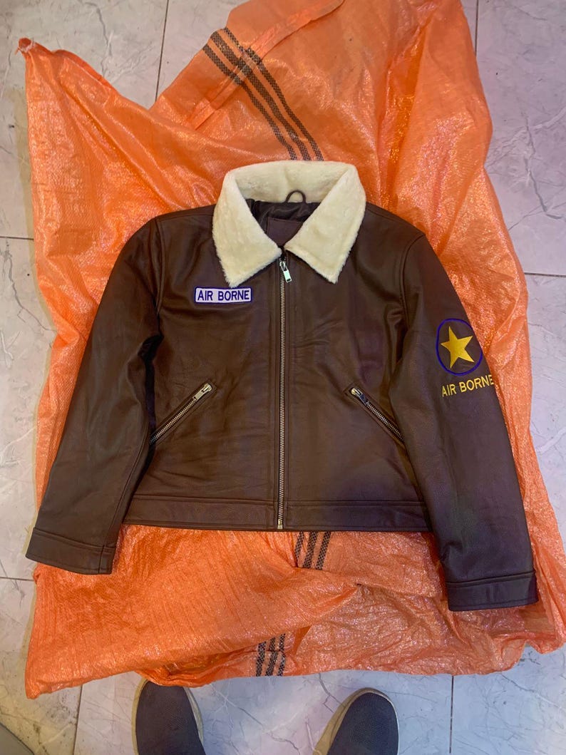 K&ouml;nnte beinhalten: Eine braune Jacke mit einem cremefarbenen Webpelzkragen und einem durchgehenden Frontrei&szlig;verschluss. Auf der Brust befindet sich ein Aufn&auml;her mit dem Text AIR BORNE, und am linken &Auml;rmel ist ein gelbes Sternemblem &uuml;ber dem Text AIR BORNE zu sehen.