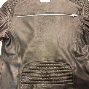 K&ouml;nnte beinhalten: Eine schwarze Lederjacke mit einem horizontalen silbernen Rei&szlig;verschluss am oberen R&uuml;cken, gesteppten Rippendetails an den Schultern und am unteren R&uuml;ckenbereich sowie einem wei&szlig;en rechteckigen Markenetikett im Nackenbereich.