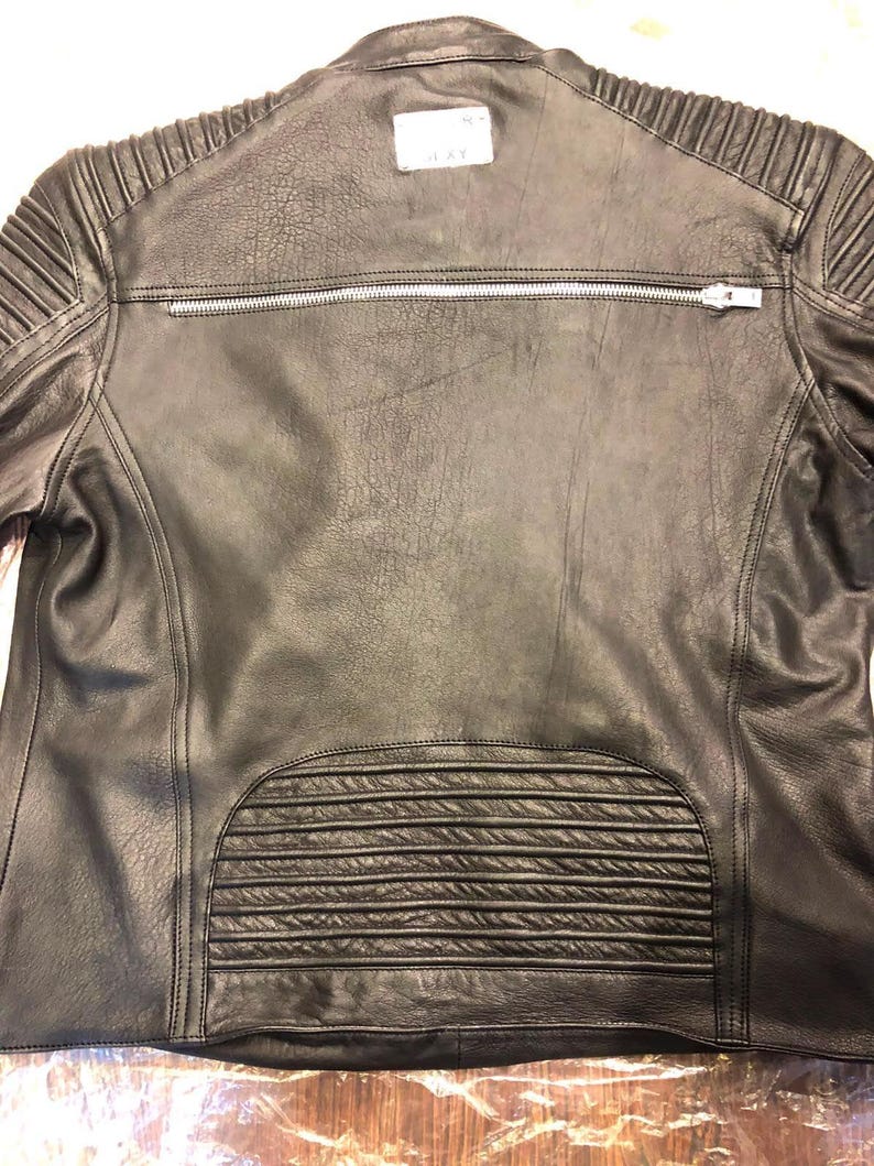 K&ouml;nnte beinhalten: Eine schwarze Lederjacke mit einem horizontalen silbernen Rei&szlig;verschluss am oberen R&uuml;cken, gesteppten Rippendetails an den Schultern und am unteren R&uuml;ckenbereich sowie einem wei&szlig;en rechteckigen Markenetikett im Nackenbereich.