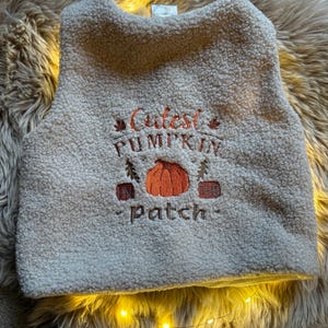 Könnte beinhalten: Eine cremefarbene ärmellose Weste aus strukturiertem Sherpa-Fleece-Stoff. Die Vorderseite zeigt ein gesticktes Design mit einer orangefarbenen Kürbisgrafik und dem Text "Cutest PUMPKIN IN THE patch" in braunem und orangefarbenem Garn.