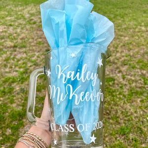 May include: A clear glass pitcher with a handle, filled with light blue tissue paper. The pitcher is decorated with white stars and the text "Kailey McKeever CLASS OF 2026".