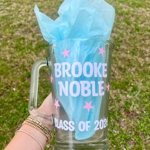 May include: A clear glass beer mug with a handle, filled with light blue tissue paper. The mug is decorated with the name "BROOKE NOBLE", pink stars, and "CLASS OF 2026" in white lettering. The mug is held against a green grassy background.