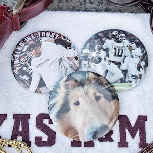 Puede incluir: Tres chapas circulares con impresiones fotográficas. Una chapa muestra a un entrenador con el texto IN ELKO WE TRUST, otra muestra a un jugador de fútbol americano con el uniforme de Texas A&M y el número 10, y la tercera muestra un perro collie.