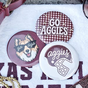 May include: Three circular pin-back buttons featuring maroon and white designs. One button has a maroon and white gingham pattern with the text GO AGGIES. Another shows a dog wearing sunglasses with the text go ags. The third features a football helmet with the text aggies.
