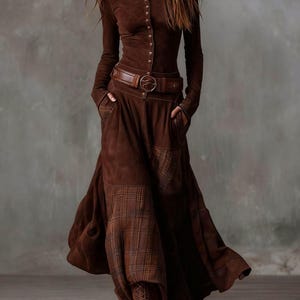 May include: A brown suede-like dress with a long-sleeved button-up top and a flowing skirt. The dress features a brown leather belt with a circular buckle and pockets. The skirt has plaid panels. Brown lace-up boots complete the outfit.