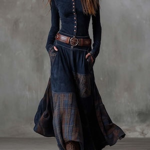 Puede incluir: Vestido azul marino de manga larga con botones frontales y cuello alto. Cuenta con un cinturón de cuero marrón y una falda larga con parches de cuadros. La modelo lleva botas marrones.