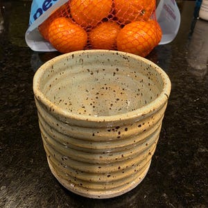 Può includere: Una ciotola in ceramica fatta a mano con una glassa beige chiaro e macchie scure. La ciotola ha un esterno a coste e una forma arrotondata. Un sacchetto di arance in una rete è sullo sfondo.