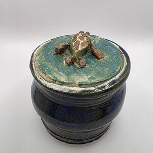 Puede incluir: Un tarro de cerámica redondo con un acabado esmaltado azul oscuro y una tapa a juego. La tapa presenta una superficie texturizada de color verde moteado con una figura de tortuga tridimensional esculpida en marrón y beige en el centro.