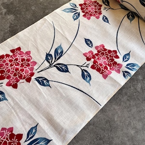 Puede incluir: Rollo de tela color crema con un estampado floral. El diseño presenta racimos de flores rojas y hojas azules sobre finos tallos azul oscuro. La tela está parcialmente enrollada, revelando el patrón.