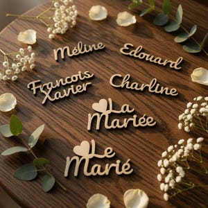 Op de afbeelding: Houten naamlabels en titels, waaronder "Meline", "Edouard", "Francois Xavier", "Charline", "La Mariée" en "Le Marié", zijn gerangschikt op een donker houten oppervlak. Witte bloemen en bloemblaadjes omringen de labels, wat een decoratief bruiloftsthema creëert.