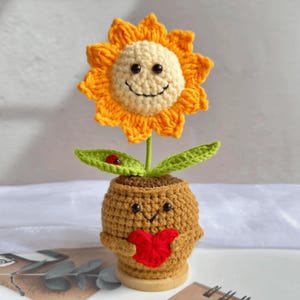 May include: A handcrafted crochet sunflower with a smiling face, orange petals, and a cream-coloured centre. The flower sits in a brown pot with a red heart, green leaves, and a ladybug. The pot has a smiling face.