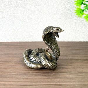 Puede incluir: Una pequeña figura de cobra de metal color bronce con el cuerpo enroscado y la capucha erguida y desplegada. La serpiente se muestra con la boca abierta, revelando colmillos detallados y escamas en toda su superficie con acabado texturizado.