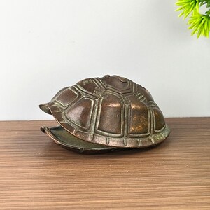 以下が含まれることがあります： ヒンジ付きの蓋が付いたブロンズ調の金属製カメの甲羅の置物。甲羅の表面にはセグメント化された幾何学模様が施されています。金属は使い古された暗い緑青仕上げで、蓋は少し開いており、中が空洞になっている様子が見えます。