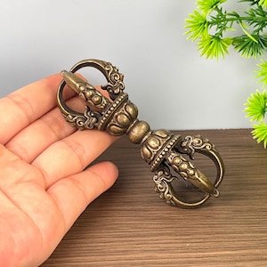 Peut inclure: Un objet rituel vajra en métal de couleur bronze avec des détails en relief complexes, une poignée centrale en forme de lotus et des griffes symétriques aux deux extrémités qui se courbent vers l'intérieur pour former une pointe.
