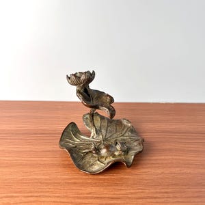 May include: A metallic bronze-toned incense holder featuring a sculpted lotus flower and a small frog resting on a large, textured lily pad base. The object has a weathered, dark patina finish with visible veins on the leaf and detailed petals.