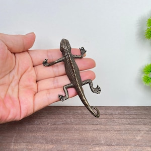May include: A bronze-toned metal lizard figurine featuring a textured, scaled surface across its back and limbs. The lizard is depicted in a crawling position with four legs extended and a long, slender tail that curves slightly at the end.