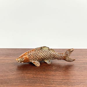May include: A metallic fish figurine with a textured scale pattern and a bronze-toned finish. The sculpture features detailed fins, a defined tail, and a realistic head shape, capturing the form of a carp in a stationary, horizontal position.