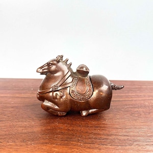 Puede incluir: Figura de caballo de color bronce con una silla de montar. El caballo está en posición de descanso, con las patas recogidas bajo el cuerpo. La silla de montar tiene detalles tallados. La figura está sobre una superficie de madera.