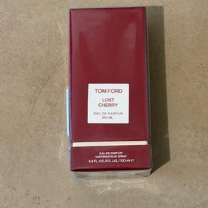 Op de afbeelding: Een rechthoekige, bordeauxrode parfumdoos met witte tekst "TOM FORD LOST CHERRY". De doos vermeldt ook "EAU DE PARFUM", "100 ML" en "3.4 FL OZ/OZ LIQ/100 ml e".