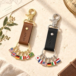 May include: Two leather keychains with gold and silver-toned metal hardware. One brown keychain is embossed with the name ATLAS and features five small flag charms. One black keychain is embossed with the name NOVA and features five small flag charms.