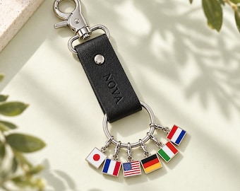 Op de afbeelding: Een zwarte leren sleutelhanger met een zilverkleurige karabijnhaak en een rechthoekige band met de inscriptie NOVA. Aan een zilverkleurige ring hangen zes kleine, vierkante, geëmailleerde bedels, elk met een afbeelding van een nationale vlag.