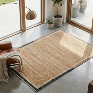 Puede incluir: Una alfombra rectangular de yute tejido de color beige natural con un borde negro. La alfombra tiene una superficie texturizada y está colocada sobre un suelo de hormigón. Es un artículo de decoración del hogar.