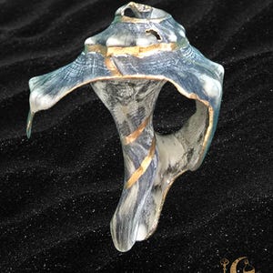 Puede incluir: Una escultura abstracta única con una forma fluida y orgánica. La pieza presenta una mezcla de tonos azules, grises y blancos, acentuados con detalles dorados. La escultura tiene una apariencia texturizada y está sobre un fondo oscuro. El logotipo "NYX & NEKTAR" es visible.