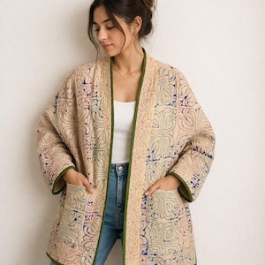 Puede incluir: Chaqueta beige con intrincados patrones bordados en rojo, azul y crema. La chaqueta tiene un ribete verde, dos bolsillos delanteros y un corte holgado. La chaqueta está abierta por delante.