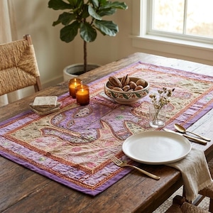 May include: A rectangular table runner featuring a patchwork design in shades of purple, tan, and orange with intricate embroidery and stitched patterns. The runner is laid flat on a wooden table, serving as a base for a ceramic bowl, candles, and plates.