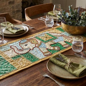 May include: A rectangular table runner featuring a central embroidered elephant motif in shades of light blue, green, and gold. The runner has a quilted texture with decorative borders and geometric patterns, displayed on a wooden dining table.