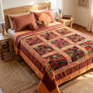Könnte beinhalten: Eine Tagesdecke mit einem Patchwork-Design aus Quadraten in Rot-, Grün- und Schwarztönen, umrandet von einem cremefarbenen Band mit einem sich wiederholenden Muster. Das Bett umfasst zwei rostfarbene Kissen und ein passendes zylindrisches Nackenkissen.