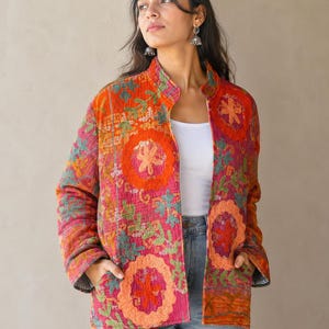May include: A quilted jacket featuring a vibrant patchwork of pink, orange, and green floral patterns with large circular embroidered motifs. The jacket has a high collar, long sleeves, and two front pockets, worn over a white tank top and blue jeans.