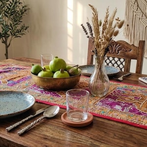 Könnte beinhalten: Ein hölzerner Esstisch mit einem leuchtend roten und violetten bestickten Tischläufer mit Elefantenmotiven, einer Messingschale mit grünen Früchten, einer klaren Glasvase mit Trockenblumen, Tellern, Besteck und Trinkgläsern.