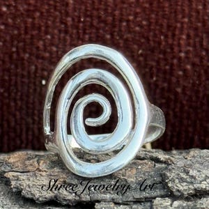 May include: A silver ring with a spiral design. The ring has a central, tightly wound spiral that expands outwards. The ring is set on a piece of wood. The text "Shree Jewelry Art" is at the bottom.