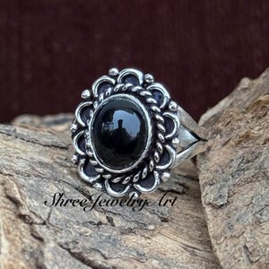 May include: A silver ring with an oval, black gemstone center. The gemstone is surrounded by a decorative, scalloped border. The ring is set on a textured wooden surface. The text "Shree Jewelry Art" is visible on the surface.