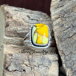 May include: A silver ring featuring a large, rectangular, yellow and green stone with a marbled effect. The ring is displayed on wood. The text "Shree Jewelry Art" is visible. The ring is a piece of jewellery.