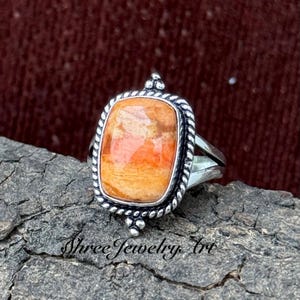 May include: A silver ring with an orange, rectangular gemstone. The stone has a textured surface and is framed by a rope-style border. The ring's band is split, and the design includes small decorative elements. The ring is a piece of jewellery.