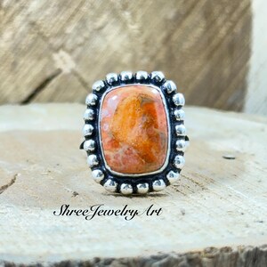 May include: A silver ring with a rectangular orange gemstone, surrounded by small silver beads. The ring is set on a wooden surface. The text "Shree Jewelry Art" is printed on the surface.