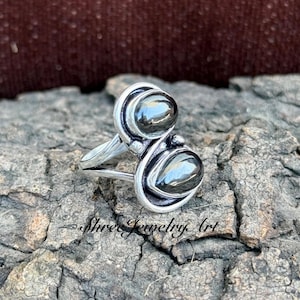 May include: A silver ring featuring two dark, oval-shaped stones. The stones are set in a decorative silver setting. The ring is displayed on a textured, gray surface. The text "Shree Jewelry Art" is visible.