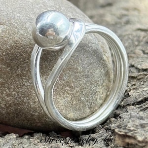 May include: A polished silver ring featuring a smooth, spherical ball accent attached to a continuous, flowing band that wraps around to form a circular shape. The metal surface is reflective, showing a clear metallic sheen and a rounded design.