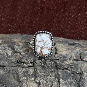 May include: A silver ring featuring a rectangular, white and brown stone. The stone is surrounded by a beaded border. The ring is set on a textured, dark surface. The text "Shree Jewelry Art" is visible.