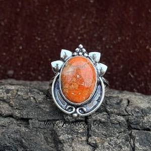 May include: A silver ring featuring an oval, orange gemstone with a speckled pattern. The ring has a decorative setting with floral-like accents and the text "Shree Jewelry Art" at the bottom.