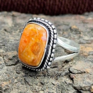 May include: A silver ring featuring a rectangular, orange-toned stone set in a bezel with a twisted rope-style border. The band is split into two separate silver shanks that connect at the base, showcasing a polished metallic finish throughout.