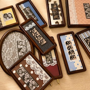 May include: A collection of dark wood picture frames featuring various photo strips and decorative backgrounds. The frames include patterns like polka dots, stripes, plaid, and cow print, accented with white lace trim and containing black and white photo strips.