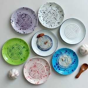May include: A collection of colorful ceramic garlic graters in various colors, including purple, green, pink, blue, and white, each with a spiral grating surface. The image also includes whole garlic bulbs and a small wooden spoon.