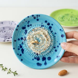 Może przedstawiać: Zestaw trzech ceramicznych talerzy do tarcia w kolorze niebieskim, fioletowym i zielonym. Niebieski talerz, trzymany w dłoni, ma starte jedzenie i ciemnoniebieskie plamki. Fioletowe i zielone talerze są w tle. Widoczny jest również migdał i gałązka tymianku.