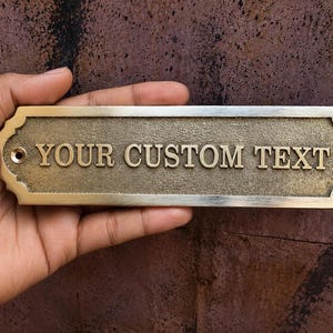 May include: A rectangular brass plaque with a textured center and smooth, scalloped edges. The plaque features the raised, embossed text "YOUR CUSTOM TEXT" in a serif font. There are two circular mounting holes, one on each far left and right side.