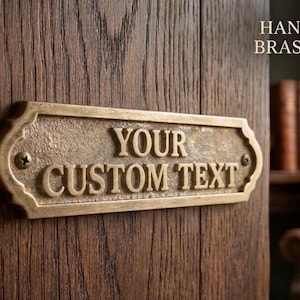 May include: A rectangular hand-cast brass plaque with decorative edges, featuring the raised text YOUR CUSTOM TEXT. The plaque is secured with two visible screws and has a textured, aged metallic finish, mounted on a dark wood surface.